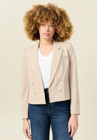 Blazer cropped beige con design doppiopetto, due bottoni e una tasca sul petto, abbinato a un top bianco a scollo a V e jeans blu.