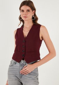 Burgundská bezrukávová vestu s výstrihom do V, piatimi čiernymi gombíkmi a dvomi prednými vreckami. Vyrobená z textúrovanej látky, skrátená pri páse.