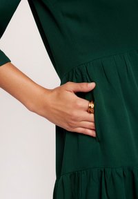 Ciemnozielona sukienka z teksturowanym korpusem i marszczoną spódnicą. Na zdjęciu widać rękę odpoczywającą na bocznej kieszeni, z złotym pierścionkiem na palcu.