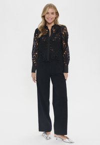 Black lace blouse with intricate cut-out patterns, long sleeves, and a buttoned front, paired with high-waisted black trousers and silver heels.