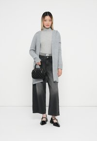 Ženska nosi svetlo sivi pulover s turtleneck ovratom, dolg sivi kardigan, črne široke hlače, črne petke in drži malo črno torbico s kratkimi ročaji.