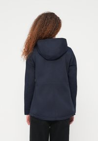 Personne aux cheveux bouclés portant une veste à capuche bleu marine et un pantalon noir, vue de dos sur un fond blanc uni.