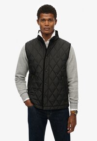 Chaleco negro acolchado con cuello alto, botones a presión en el frente y bolsillos laterales con cremallera; llevado sobre un suéter gris y jeans oscuros.