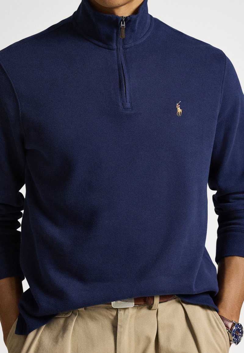 Man wearing navy blue quarter-zip sweater with embroidered polo player logo, beige pleated trousers, brown belt, and blue wristwatch.