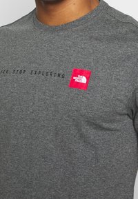 Šedé bavlněné tričko s kulatým výstřihem, krátkými rukávy a červeným obdélníkovým logem s textem "THE NORTH FACE" a "NEVER STOP EXPLORING."