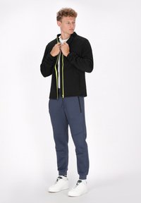 Giacca nera con zip, dotata di una cerniera gialla neon, abbinata a pantaloni della tuta blu e sneakers bianche. Caratterizzata da un colletto rialzato e tasche laterali.