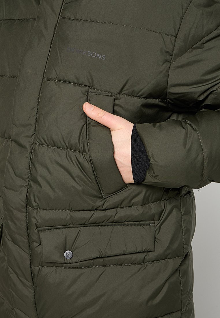 Hand in der Tasche einer olivgrünen gepolsterten Jacke mit gesteppten Details, Klappentaschen und sichtbar aufgesticktem Markenlogo auf der Brust.
