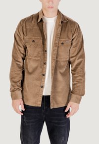 Camicetta in corduroy marrone con due tasche sul petto, chiusura con bottoni e tessuto testurizzato. Indossata sopra un maglione bianco a coste e jeans scuri.