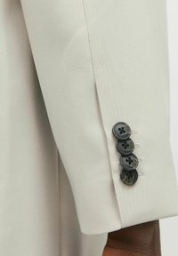 Manga de una chaqueta entallada de color gris claro con cuatro botones negros de efecto marmoleado, costuras detalladas y una textura de tela suave.