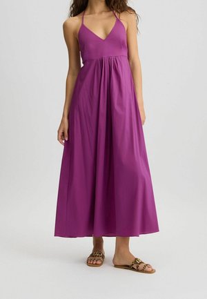 Kvinde iført en ærmeløs lilla maxikjole med dyb V-hals og leopardmønstrede flade sandaler, stående mod en ensfarvet baggrund.