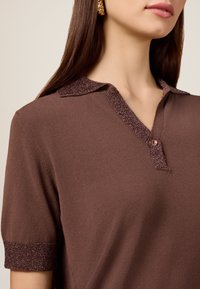 Brun polo-skjorte med en tekstureret krave og manchetter med metalliske detaljer, knaplukning og et glat strikstof.