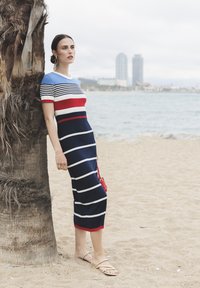 Randig stickad klänning med korta ärmar, med blå, röda och vita horisontella ränder. Halvlång med en åtsittande passform. Guld sandaler och en röd väska.