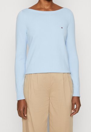 Person trägt einen hellblauen Pullover mit langen Ärmeln und kleinem Logo auf der Brust sowie beige plissierte Hosen und steht vor einem weißen Hintergrund.