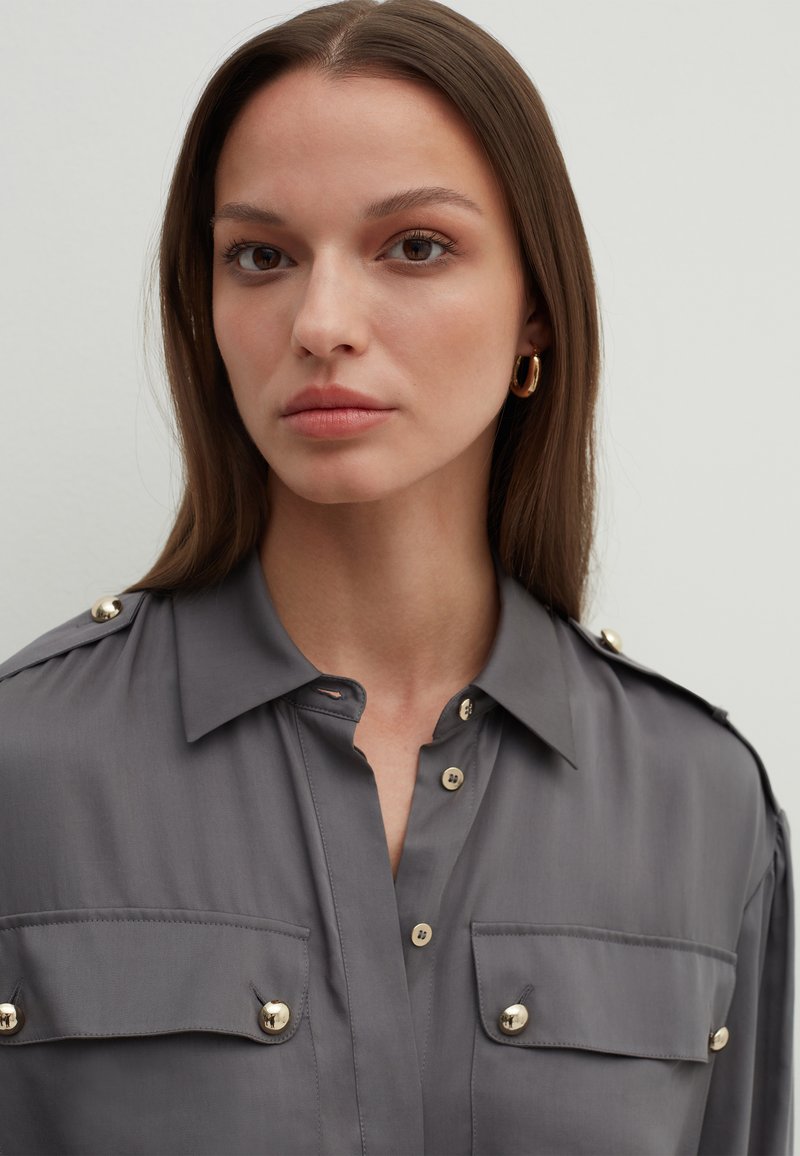 Giovane donna con lunghi capelli castani, che indossa una camicia grigia con bottoni dorati e orecchini a cerchio dorati, espressione neutra.