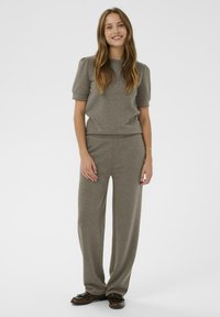 Žena stojaca a usmievajúca sa, oblečená v taupe krátkom rukávovom top s vyvýšenými ramenami, zodpovedajúcich širokých nohaviciach a tmavohnedých loaferoch.