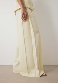 Gaiši dzelteni platli kantaini bikses, izgatavotas no mīksta, plūstoša materiāla. Aprīkotas ar izsmalcinātām krokām un vaļīgu piegriezumu, saskaņotas ar plakaniem zābakiem.