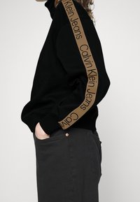 Sudadera negra con cuello alto, que presenta un logo marrón de Calvin Klein Jeans en la manga. Combinada con pantalones gris oscuro.