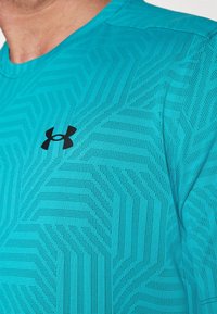 Turkos träningsskjorta med ett strukturerat mönster, som har vinklade former och små perforeringar. Svart Under Armour-logotyp på vänster bröst.