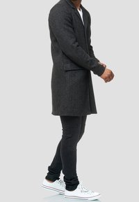 Abrigo largo de punto gris con cierre de un solo botón, bolsillos laterales y ajuste entallado, llevado sobre pantalones negros ajustados y zapatillas blancas.