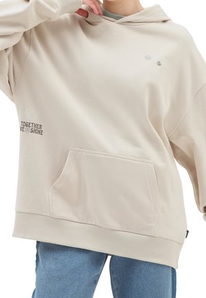 Sweat-shirt à capuche beige en matériau doux. Comprend une poche frontale, un ourlet côtelé et des graphiques avec l'inscription "ENSEMBLE NOUS BRILLONS" accompagnée d'un design de globe.