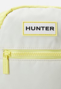 Gros plan de la poche avant d'un sac à dos jaune clair et blanc avec une fermeture éclair jaune et une étiquette de marque « HUNTER » au-dessus de la fermeture éclair.