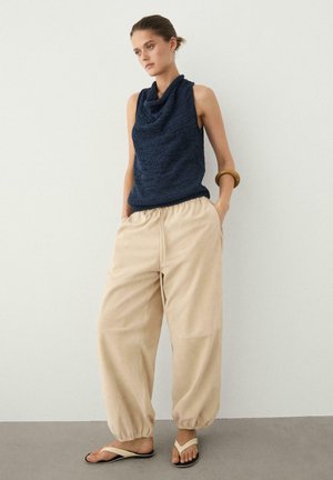 Femme debout contre un mur blanc portant un haut sans manches bleu marine à col bénitier, un pantalon beige ample avec des poignets élastiques et des tongs beiges.