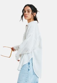 Witte, longsleeve shirt van een gestructureerde stof met een gebogen zoom, gecombineerd met een lichtblauwe denim rok. Persoon houdt een paar bruine zonnebril vast.