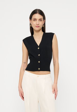 Ženska, oblečena v brezrokavnik črnega gumbastega kratkega topa in bele hlače z visokim pasom, stoji ob enobarvnem belem ozadju.