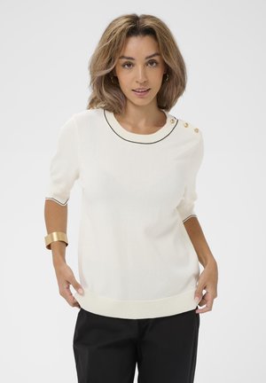 Woman with shoulder-length hair wears white short-sleeve top with gold buttons on shoulder, black pants, gold cuff bracelet, and hoop earrings.