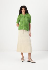 Zelený pletený krátký cardigan s knoflíkovou légou, spárovaný s krémovou sukní až ke kolenům. Modelka má na sobě světlé balerínky.