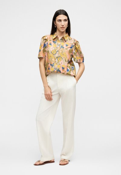 Γυναίκα που στέκεται με τα χέρια στις τσέπες, φορώντας ένα κοντομάνικο πουκάμισο με λουλούδια και κουμπιά, άσπρο παντελόνι με φαρδιά μπατζάκια και επίπεδα σανδάλια.