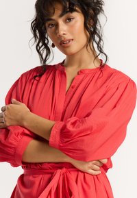 Korallefarbendes Kleid mit Knopfausschnitt, Puffärmeln und tailliertem Gürtel. Verfügt über einen strukturierten Stoff und bestickte Details an den Ärmeln.