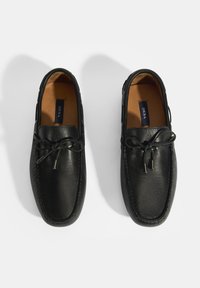 Zwarte lederen loafers met een gladde textuur, contrasterende stiksels, en een vetersluiting aan de voorkant met metallic accenten. Binnenvoering is lichtbruin.