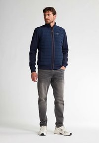 Gilet trapuntato blu navy con maniche a contrasto, cerniera frontale e toppa con logo. Jeans grigi e scarpe sportive bianche completano l'outfit.