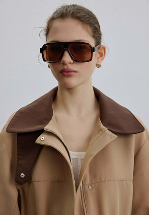 Giovane donna che indossa occhiali da sole marroni oversize e giacca beige con grande colletto marrone scuro su sfondo semplice.