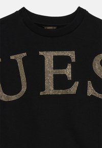 Black sweatshirt featuring large 'GUESS' lettering in gold rhinestones, round neckline, and ribbed cuffs. Soft texture with a solid color background.