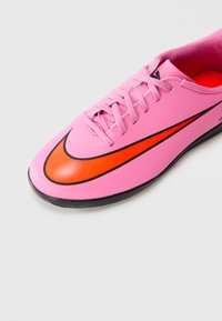 Rózsaszín sportcipő piros és fekete Nike logóval, rózsaszín fűzőkkel és fekete talppal, fehér háttéren.