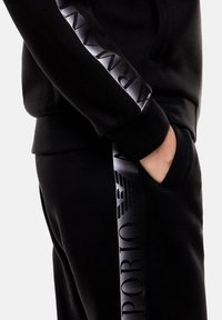 El sudadera y los pantalones negros presentan franjas negras brillantes con el texto y logotipo de "EMPORIO ARMANI". El material parece suave y texturizado.