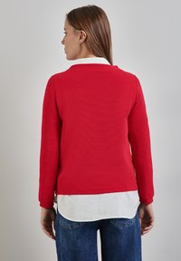 Rotes Langarmoberteil mit strukturierter Oberfläche, rundem Ausschnitt und weißem Shirt-Layer am Saum. Getragen mit dunkelblauen Jeans.