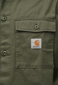 Πράσινο πουκάμισο ελιάς με μία τσέπη στο στήθος, δύο πράσινα κουμπιά και λευκή ετικέτα με το λογότυπο Carhartt με πορτοκαλί λεπτομέρεια. Ομαλό, ανθεκτικό ύφασμα.