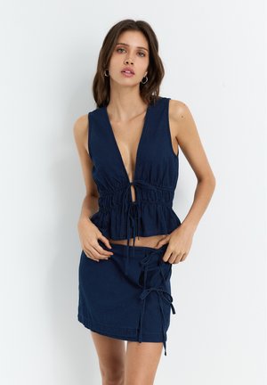 Navy sinivihreä kahden osan asu, jossa on peplum-toppia syvällä V-kaula-aukolla ja säädettävillä nauhoilla, yhdistettynä mini-hameeseen, jossa on sivusiteet.