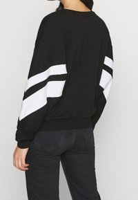 Sudadera negra con rayas diagonales blancas en las mangas; corte relajado y cuello de canalé. Combinada con vaqueros oscuros.