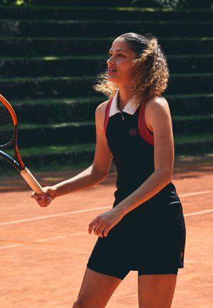 Black tennis dress with red accents, features a collar and zip, made of stretchable fabric. She holds an orange tennis racket on a clay court.