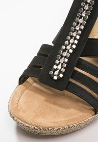 Gros plan d’une sandale noire avec une semelle intérieure beige rembourrée et des perles argentées décoratives le long de la bride.