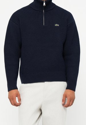 Homme portant un pull en maille texturée bleu marine avec une fermeture éclair quart et un logo crocodile vert sur la poitrine, associé à un pantalon blanc.