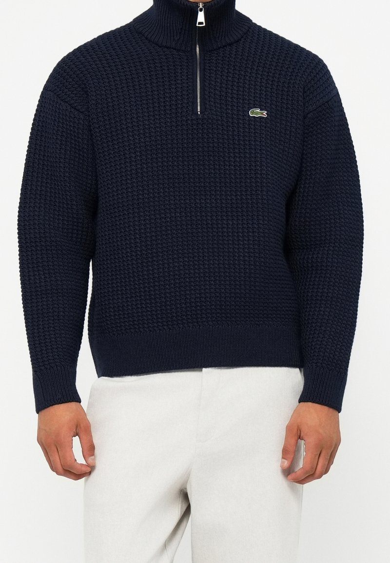 Homme portant un pull en maille texturée bleu marine avec une fermeture éclair quart et un logo crocodile vert sur la poitrine, associé à un pantalon blanc.