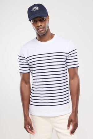 Homme portant une casquette marine, un T-shirt blanc avec des rayures horizontales marines et un pantalon de couleur claire, se tenant contre un fond blanc uni.
