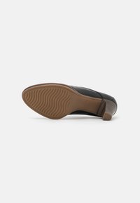 Chaussure en cuir noire unique avec semelle en caoutchouc marron et talon bas empilé, vue par le dessous sur fond blanc.