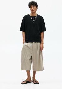 T-shirt oversize noir à manches courtes avec des détails en bouton, associé à un short à jambes larges beige et des sandales noires. Colliers superposés en argent.