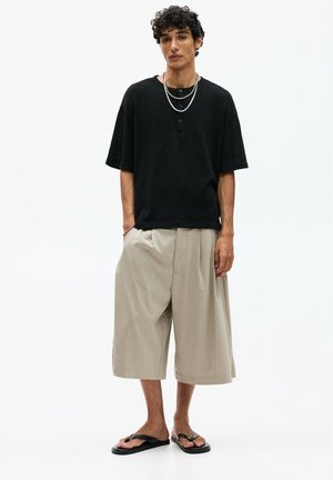 Tricou negru oversize cu mâneci scurte și detalii cu butoane, purtat cu pantaloni scurți bej cu picior larg și sandale negre. Coliere suprapuse din argint.